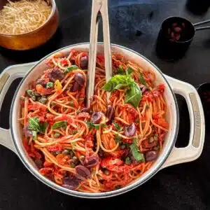 Pasta puttenesca in a Dutch oven and garnished with extra olives, basil and capers.