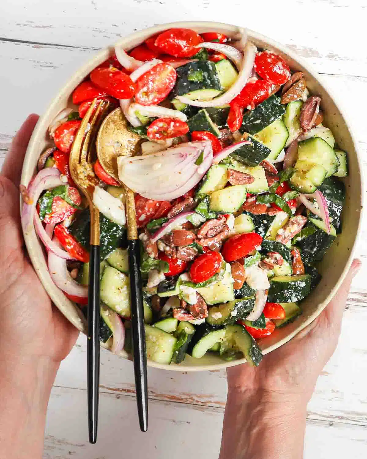 A pair of hand holding a Greek salad that has been tossed together in a bowl.