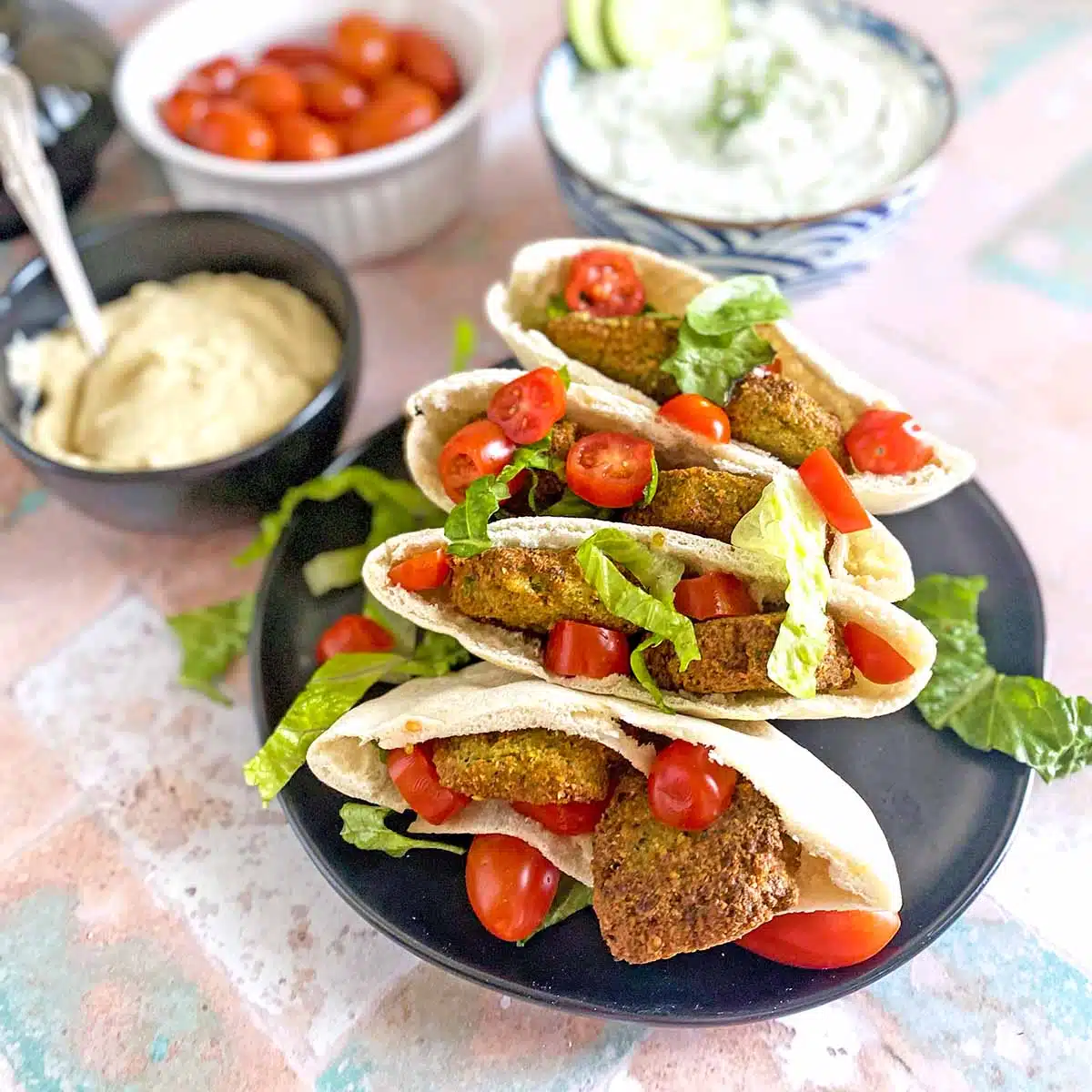 Four falafel pita sandwiches with toppings next to it. 
