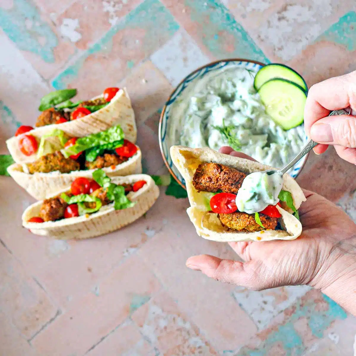 A hand holding a pita pocket with falafel patties in it.  The other hand is spooning dairy-free tzatziki into the pocket. 
