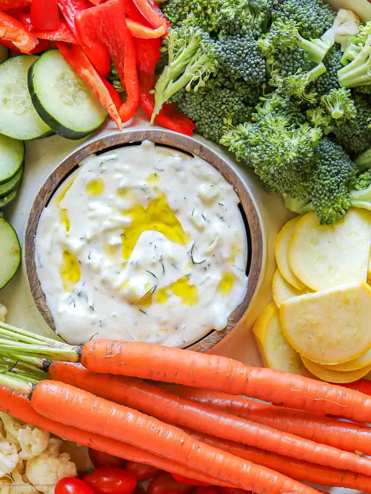 A bowl of dairy-free tzatziki surrounded by raw carrots, yellow squash slices, cauliflower florets, and tomatoes.