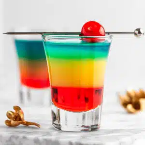 Shot glass with layered rainbow drink garnished with a cherry.