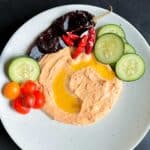 A plate filled with harissa hummus surrounded with cucumbers, tomatoes and pita bread.