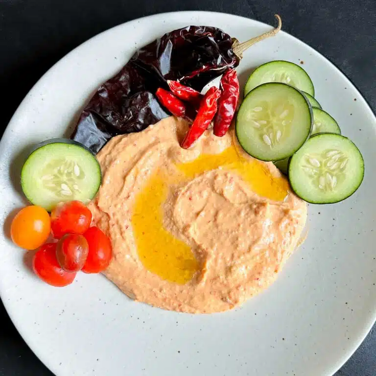 A plate filled with harissa hummus surrounded with cucumbers, tomatoes and pita bread.