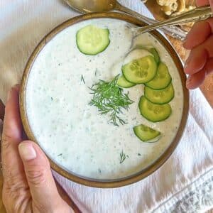 Creamy cold cucumber soup with fresh dill sprigs and cucumber slices on top and a hand holding a spoon.