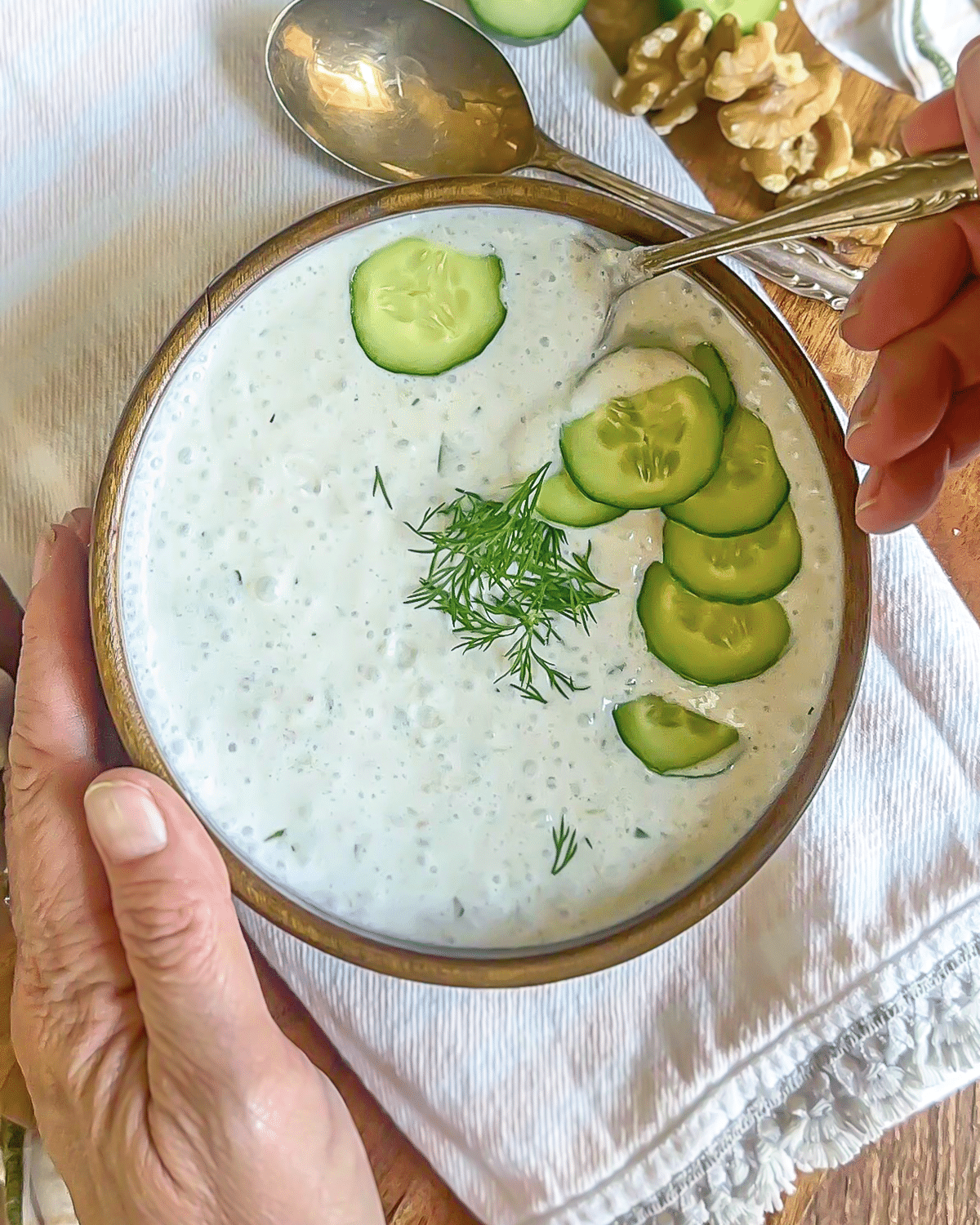 Creamy cold cucumber soup with fresh dill sprigs and cucumber slices on top and a hand holding a spoon.