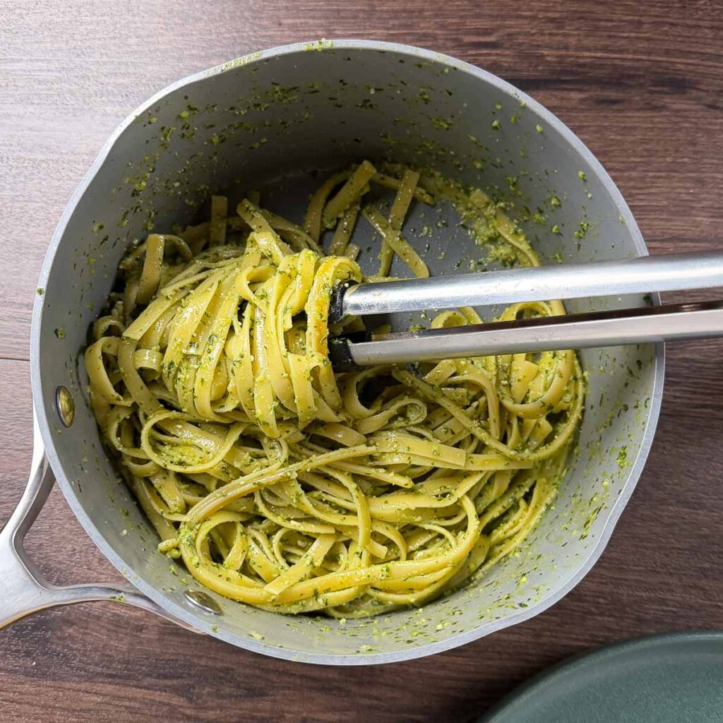 Pesto pasta in a saucepan coiled around tongs.