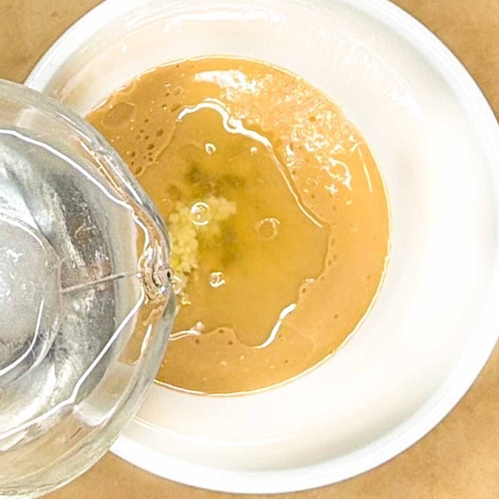 Pouring water into a bowl with tahini paste, lemon juice, minced garlic and salt to create tahini sauce.