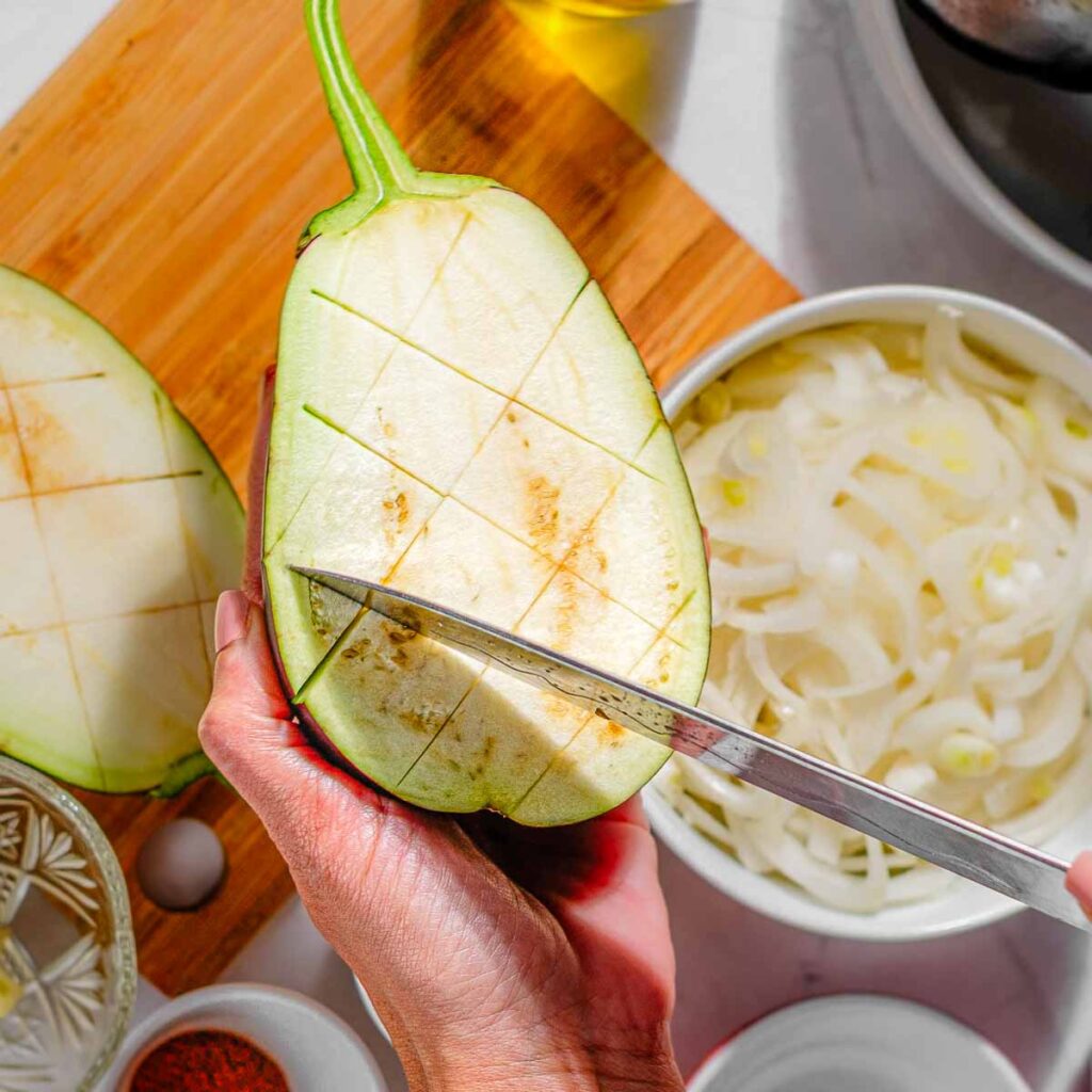 A hand holding an eggplant with deep criss-crosses cut into the vegetable.
