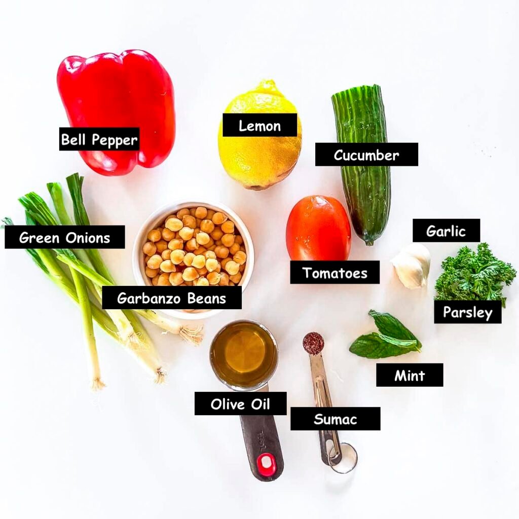 Ingredients used in making balela, a Middle Eastern salad. Ingredients include garbanzo beans, bell pepper, lemon, cucumber, tomatoes, garlic, parsley, mint, sumac, olive oil and green onions.
