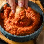 A hand dipping a chip into a green bowl filled with ajvar.