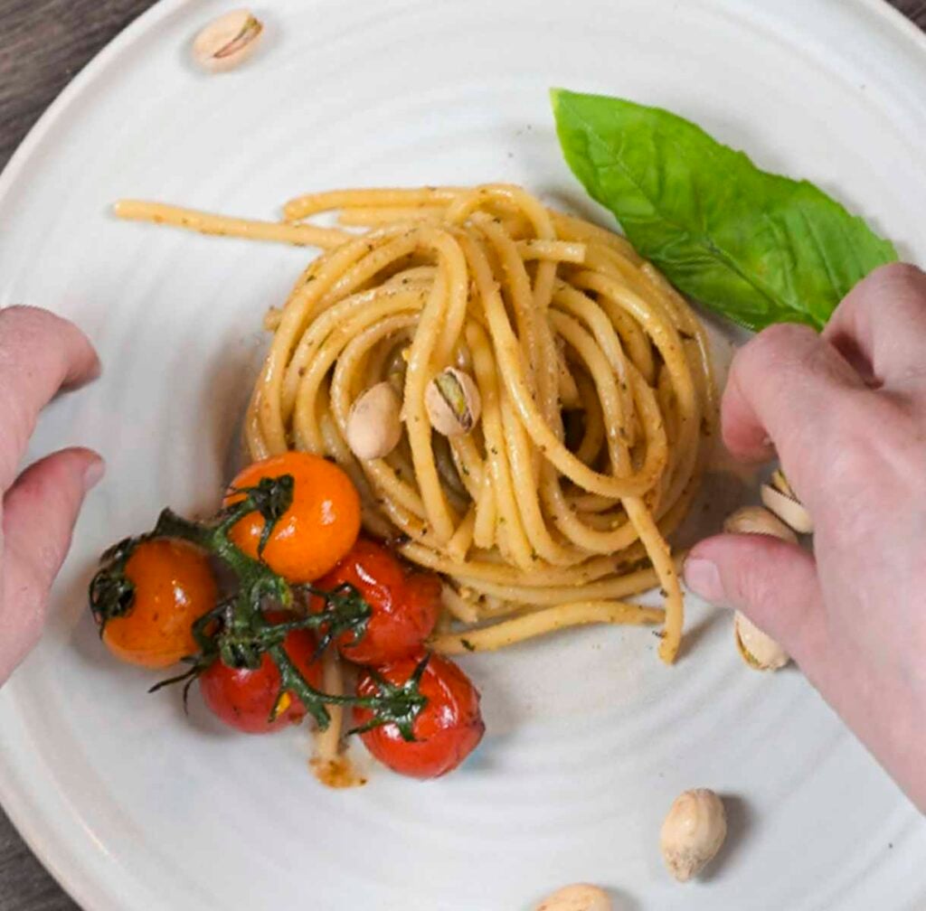 Garnishing pesto pasta with blistered tomatoes, a basil leaf and pistachio nuts in their shells.