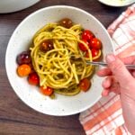 A bowl filled with pistachio pesto that has been tossed with thick pasta. A hand is holding a fork getting ready to try a bite.