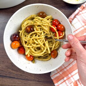 A bowl filled with pistachio pesto that has been tossed with thick pasta. A hand is holding a fork getting ready to try a bite.