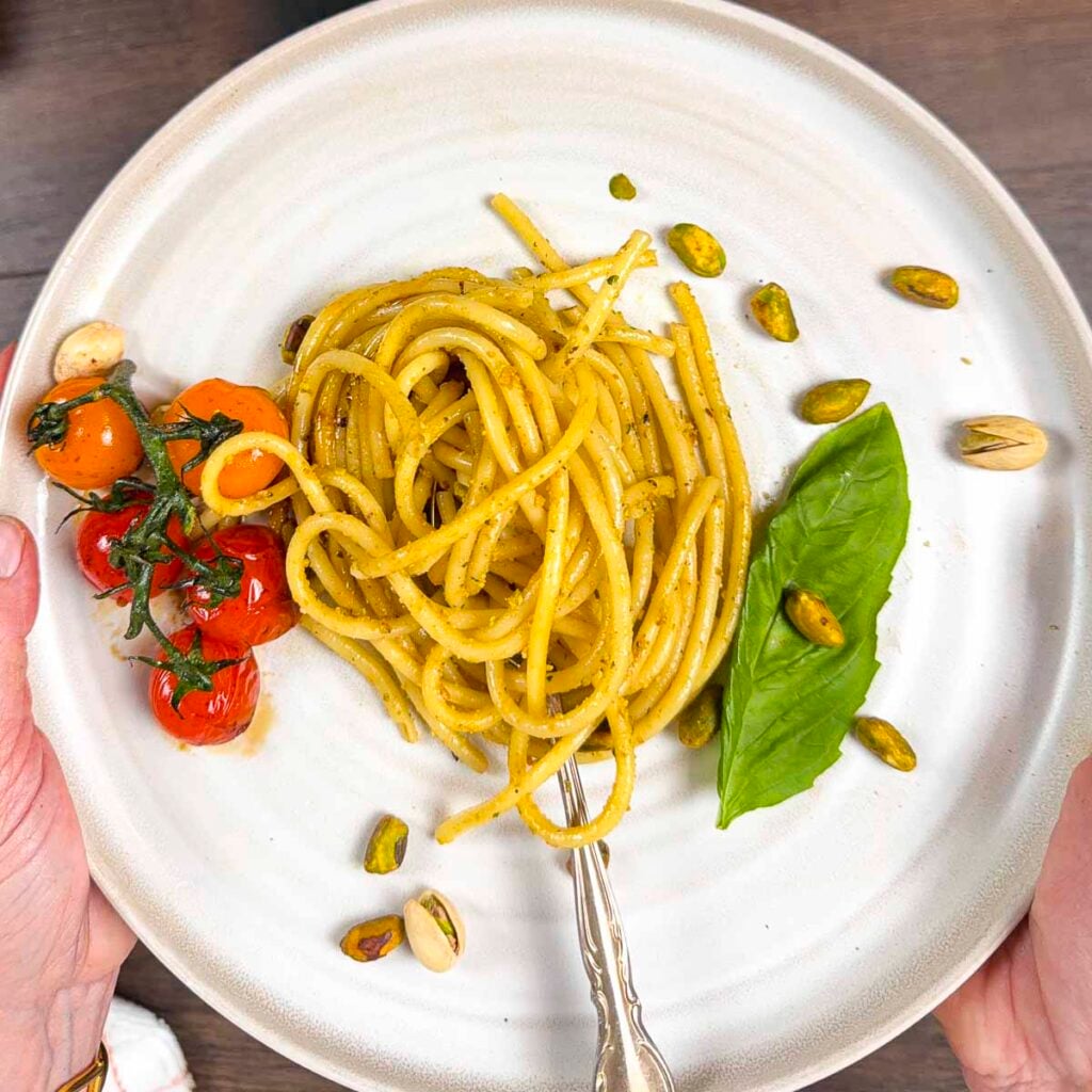 Pistachio pesto pasta wrapped into a coil with pistachios and blistered tomatoes being held up to the camera.