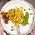 Pistachio pesto pasta wrapped into a coil with pistachios and blistered tomatoes being held up to the camera.