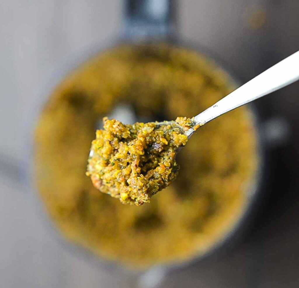 A spoon filled with pistachio pesto is held over the food processor.