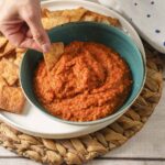 Balkin ajvar in a green bowl with a hand dipping a chip into the ajvar.