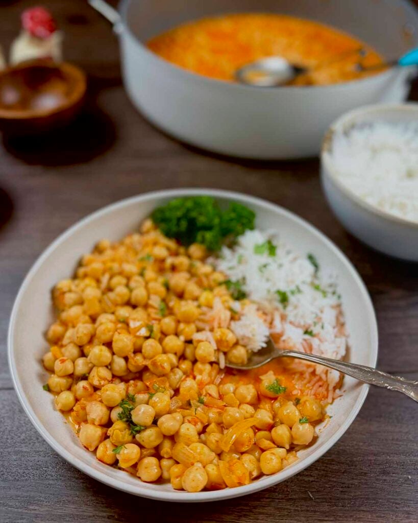 A plate filled with spicy Moroccan harissa chickpeas. The plate also has rice and is garnished with parsley and mint.