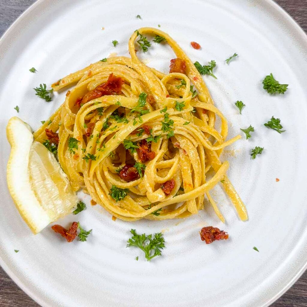 Vegan creamy pasta with sun-dried tomatoes on a white plate. The pasta has been garnished with parsley, sun-dried tomatoes, and parsley.