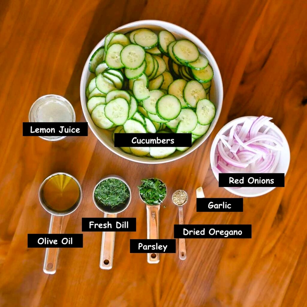 Ingredients used in making a cucumber and dill salad (cucumbers, red onion, garlic, dried oregano, parsley, fresh dill, olive oil, and lemon juice).