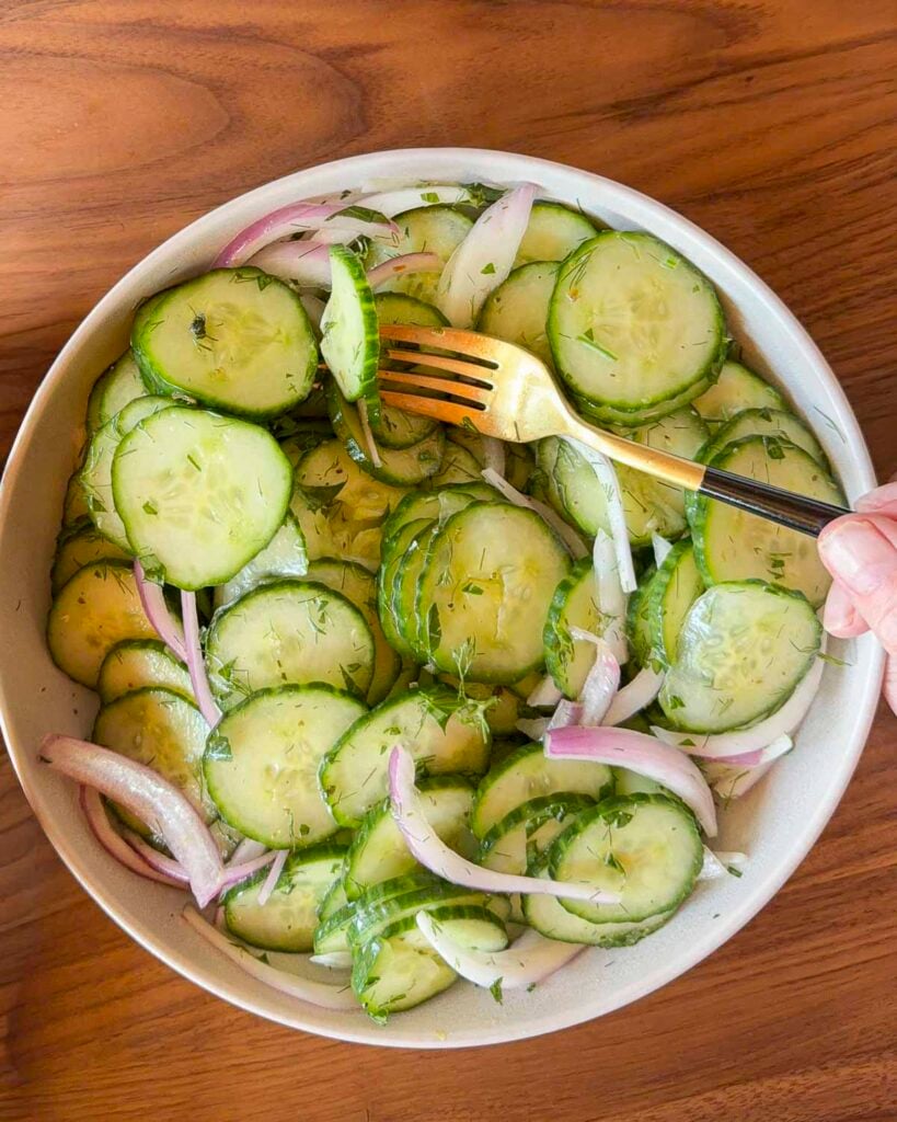 A bowl filled with sliced cucumbers, red onion and a cucumber salad dressing. There is a hand holding a fork in the bowl.