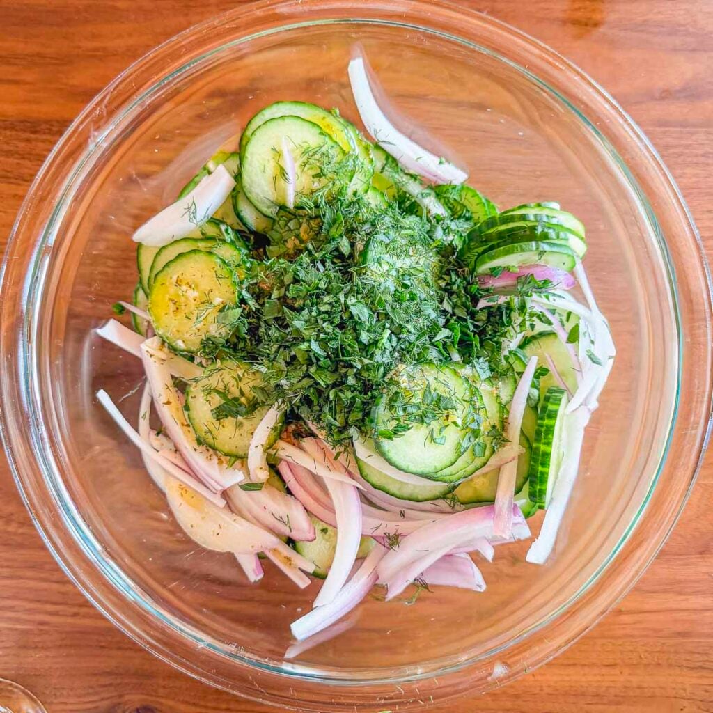 A bowl filled with sliced cucumbers, red onion, fresh dill, and parsley before being tossed into a cucumber salad.