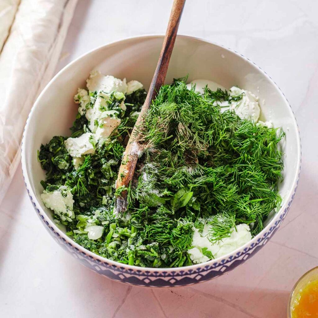 A bowl filled with the cooked spinach filling, vegan feta, plant-based yogurt, dill, and parsley.