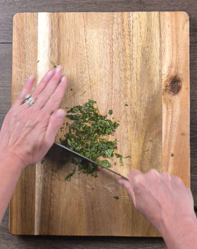 A cutting board and two hands holding a knife, chopping parsley.