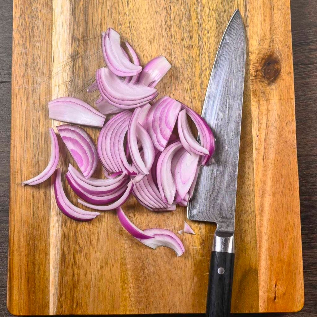 Red onions cut into thin slices on a cutting board with a chef knife next to them.