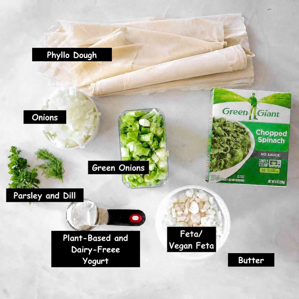 Ingredients needed for making vegan spanakopita (phyllo dough, frozen spinach, onion, green onions, parsley, dill, plant-based yogurt and vegan feta cheese.