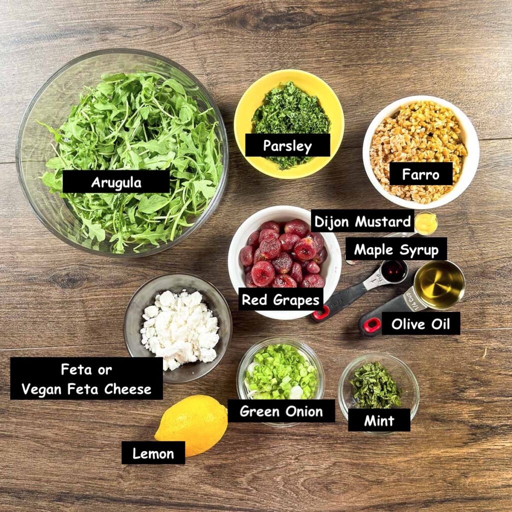 Ingredients used in making a farro grain salad (farrow, arugula, seedless red grapes, feta/vegan feta cheese, parsley, green onion, mint, lemon, olive oil, maple syrup, and Dijon mustard).