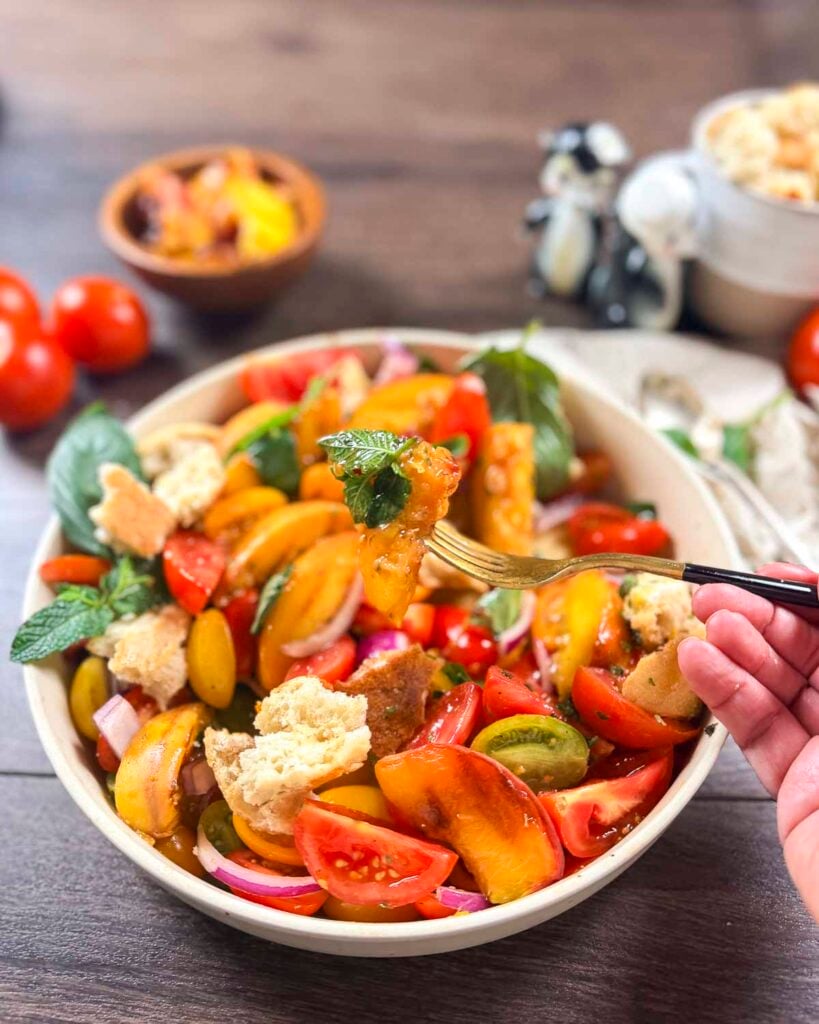 Close-up of a hand with a fork holding a grilled peach over a summer panzanella salad.