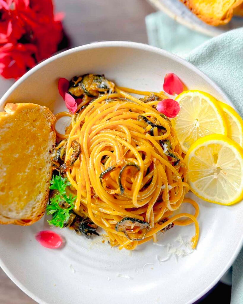 Spaghetti twirled with golden zucchini rounds on a while plate garnished with bread and lemon slices.