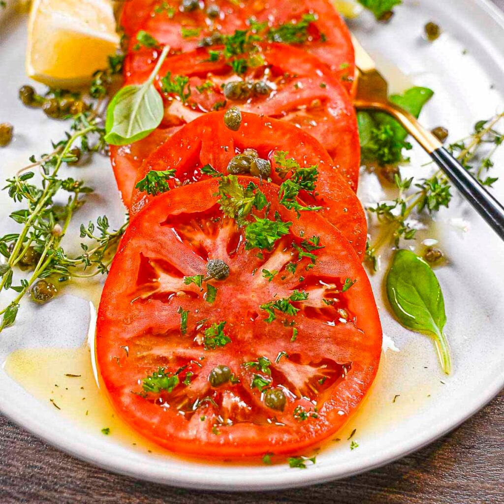 Red marinated beefsteak tomatoes with capers and fresh thyme. The first tomato slice has a bite taken out of it.