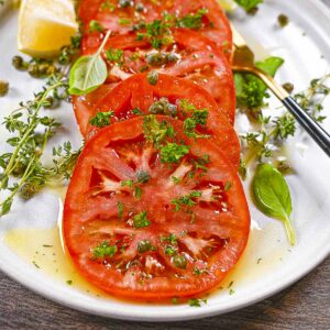 Red marinated beefsteak tomatoes with capers and fresh thyme. The first tomato slice has a bite taken out of it.