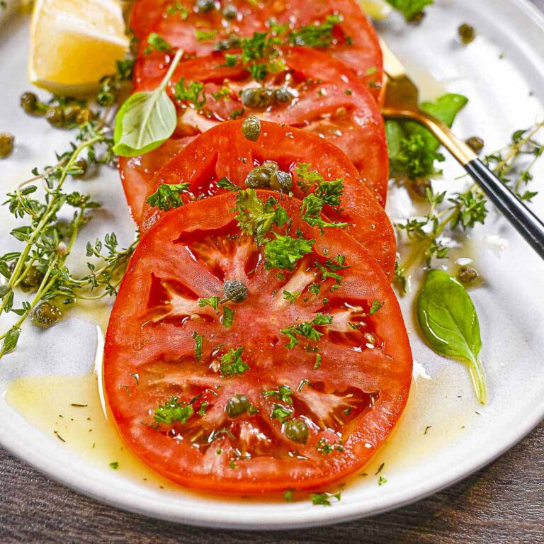 Red marinated beefsteak tomatoes with capers and fresh thyme. The first tomato slice has a bite taken out of it.