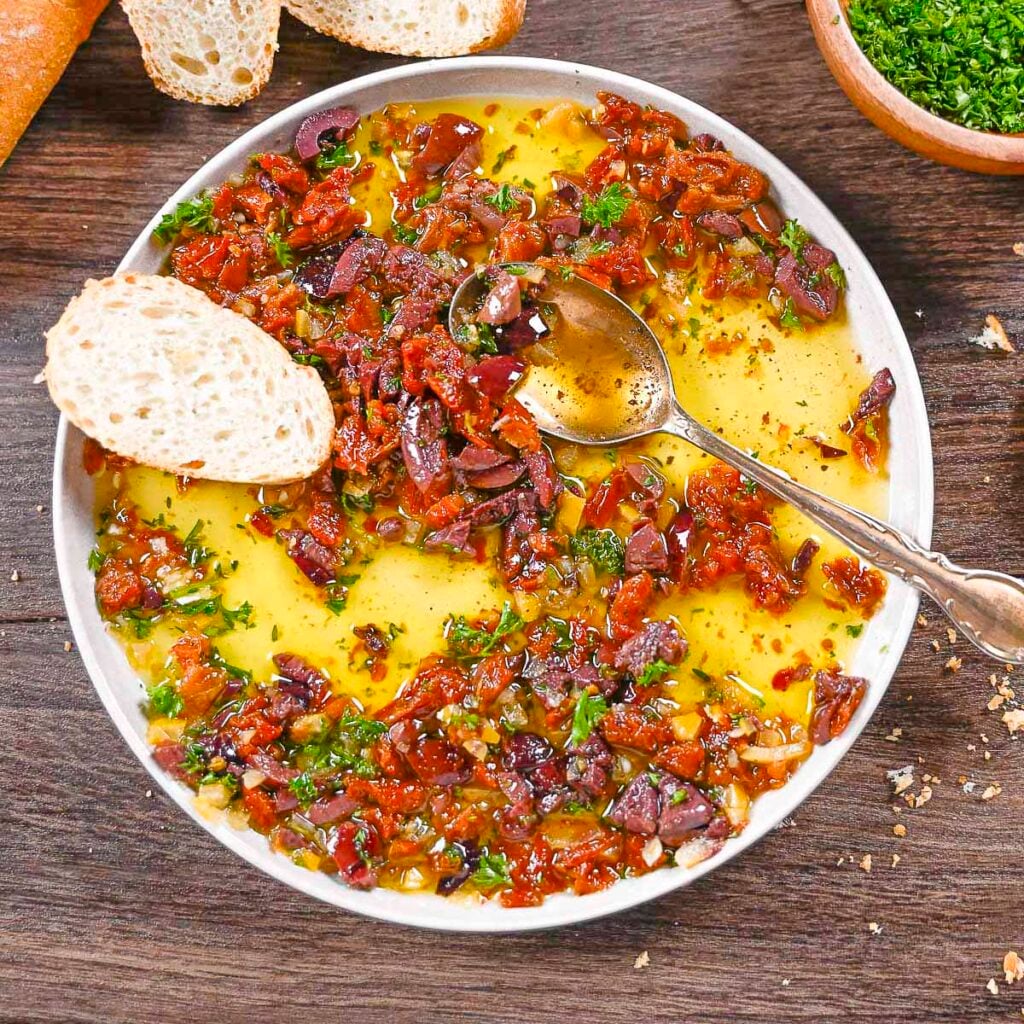 Mediterranean bread dip made with Kalamata olives, sun-dried tomatoes, and olive oil with a spoon on the plate and a toast point.