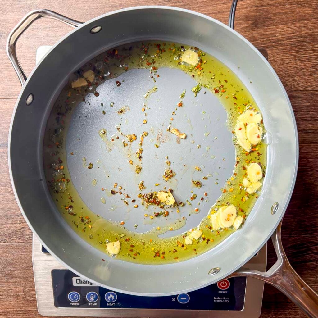 A skillet filled with olive oil, rosemary, crushed red pepper flakes, and sliced garlic.