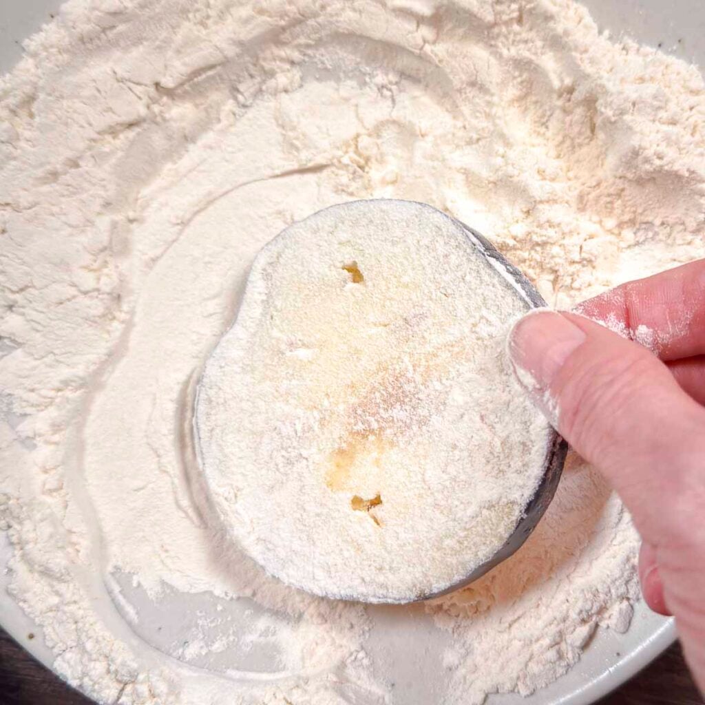 A hand dredging a slice of eggplant in flour.