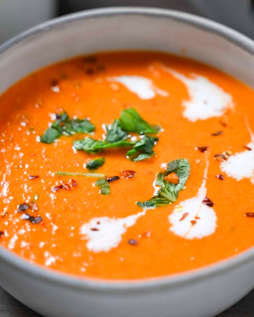 Vegan roasted red pepper tomato soup in a white bowl with torn basil leaves, red pepper flakes, and vegan heavy cream.