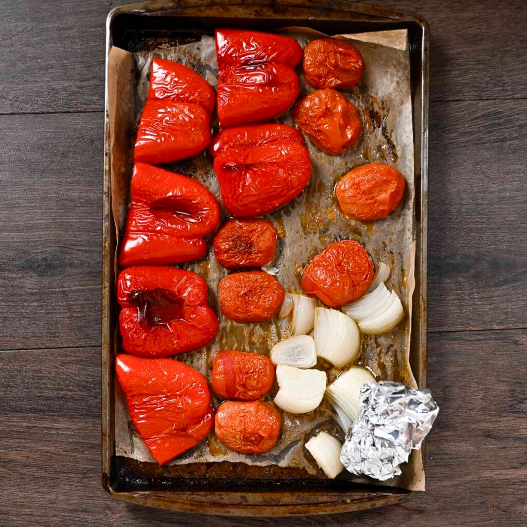 Red peppers, tomatoes, onion and a whole garlic head on a sheet pan. All of the vegetables have been oven-roasted.