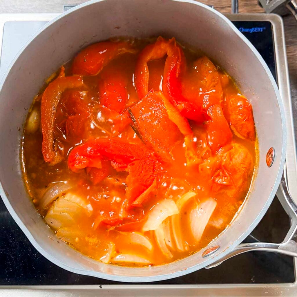 A saucepan filled with vegetable broth, red peppers, tomatoes, onion and garlic that have been oven-roasted.