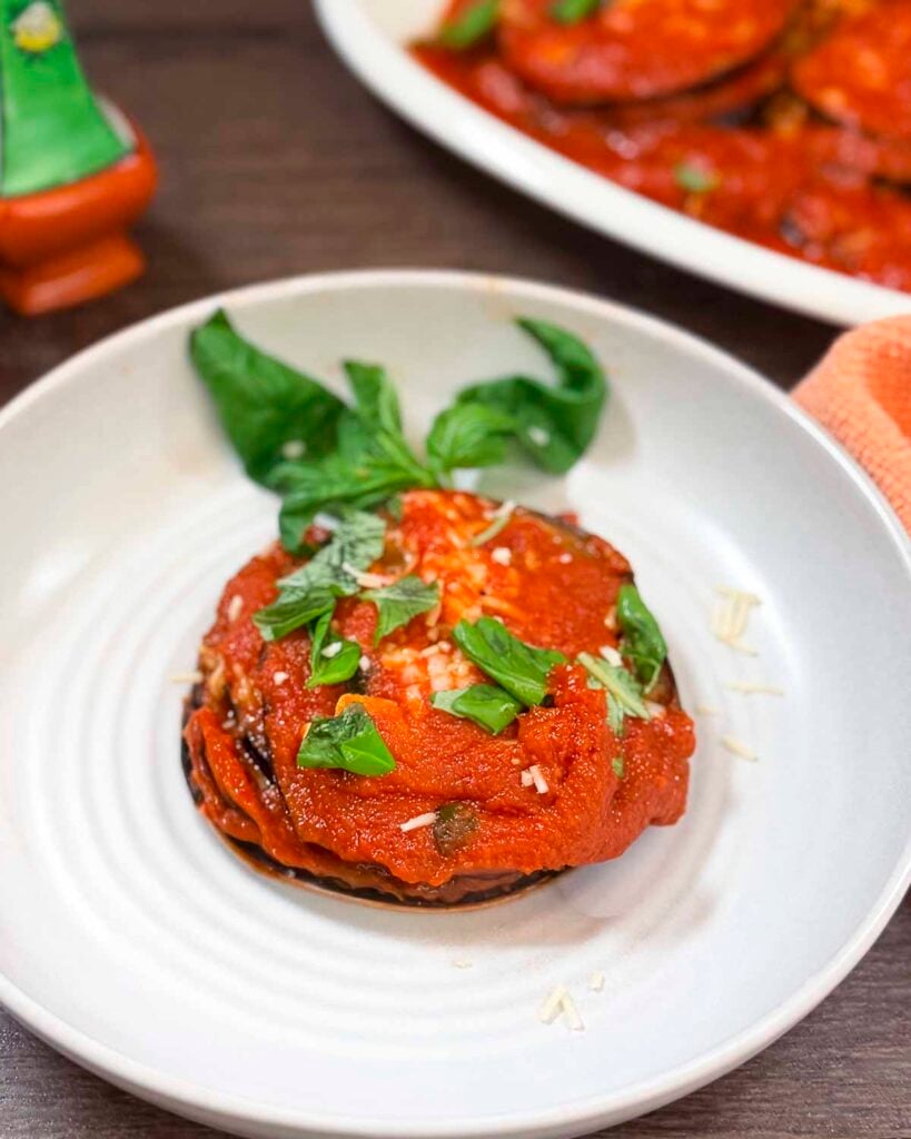 A stack of vegan eggplant parmigiana on a plate topped with freshly torn basil and vegan Parmesan.