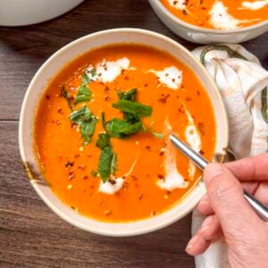 Vegan roasted red pepper tomato soup in a white bowl with torn basil leaves, red pepper flakes, and vegan heavy cream.