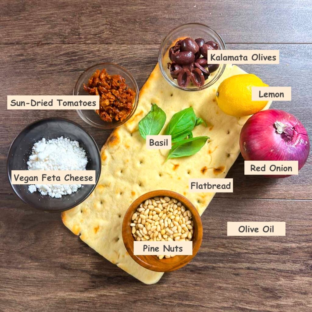 Ingredients used in making a vegan Mediterranean flatbread pizza (flatbread, olive oil, red onion, Kalamata olives, sun-dried tomatoes, vegan feta cheese, pine nuts, lemon and fresh basil leaves.)