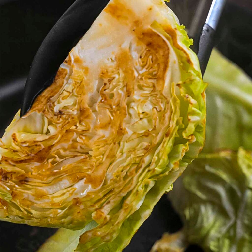 A pair of tongs holding up a seared cabbage wedge to the camera.